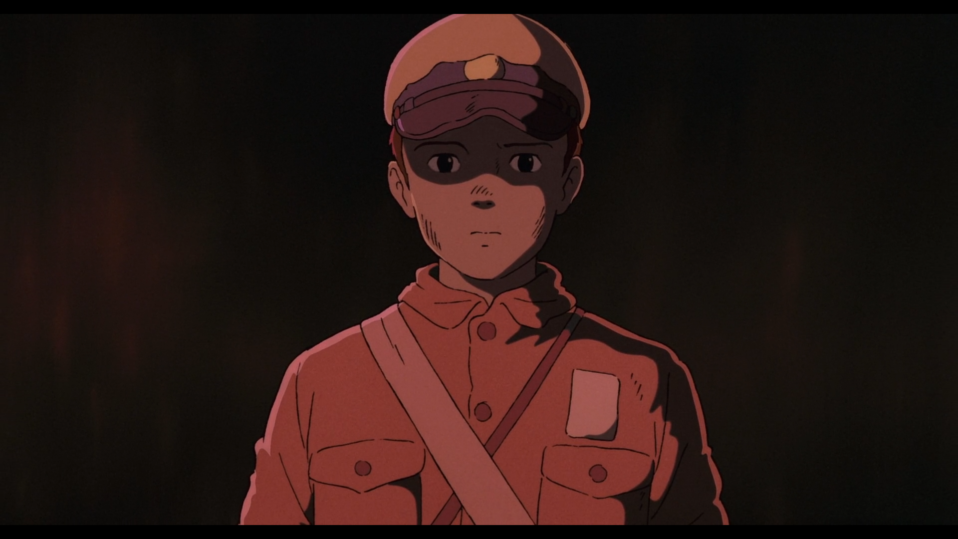 Première image du film le Tombeau des Lucioles
Seita, adolescent japonais de 14 ans, aux cheveux foncés et aux yeux noirs, portant une chemise foncée avec deux poches au niveau de la poitrine, deux sacs dont les bandoulières, une claire, une plus foncée, se croisent et une casquette blanche avec une visière foncée, regarde fixement devant lui.
Il est baigné dans une lumière rougeâtre qui rend les couleurs difficiles à distinguer.
Le fond derrière lui est noir.